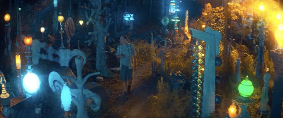 a man stands in the middle of a sculptural garden that he created himself, with lighting features that glow in blues and yellows