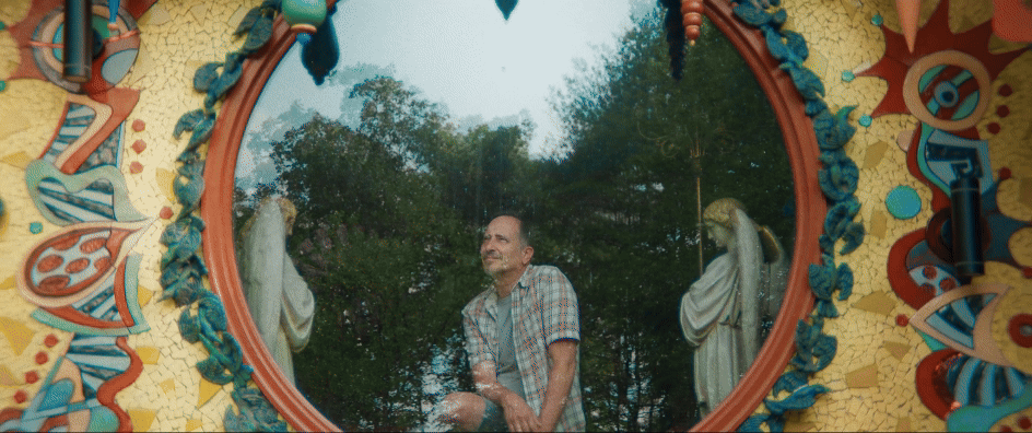 a gif from a short documentary of an artist seated inside of a round window in a colorful, whimsical house he built himself