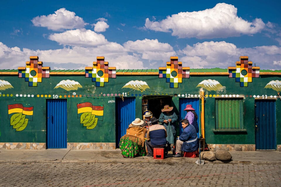 a group of people gather in front of a colorful, mostly green building set against a blue sky with puffy clouds