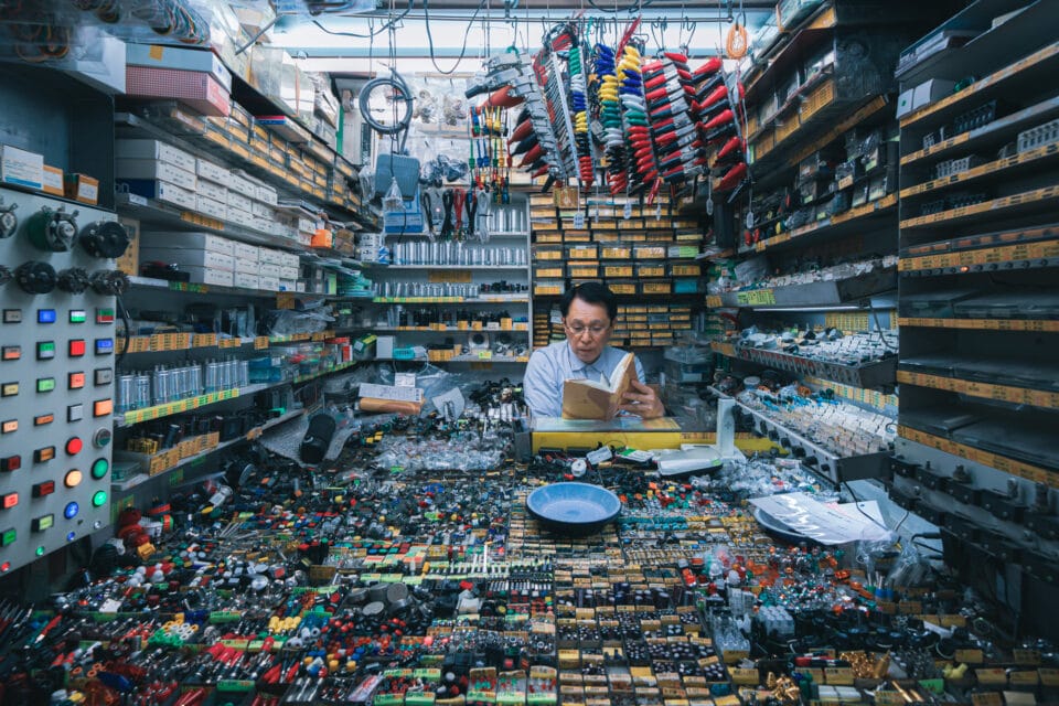 a man sits inside of a visually overwhelming selection of electronics and items available for purchase