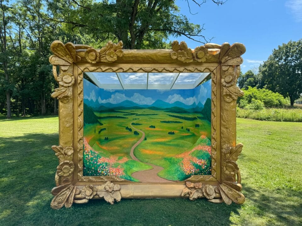 a very large, "walk-in" painting situated in a grassy environment. the installation consists of a massive ornate gold frame, and a hilly green landscape inside.