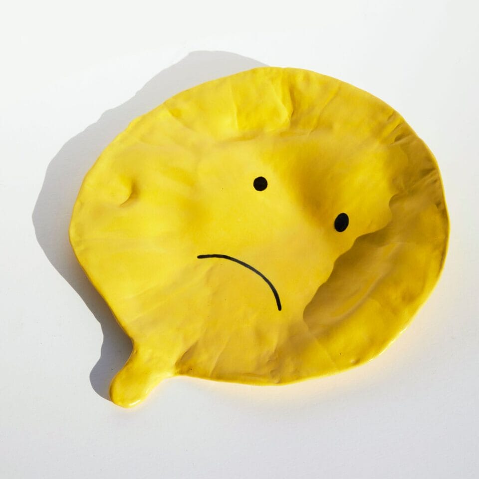 a ceramic sculpture of a deflated yellow balloon with a sad face