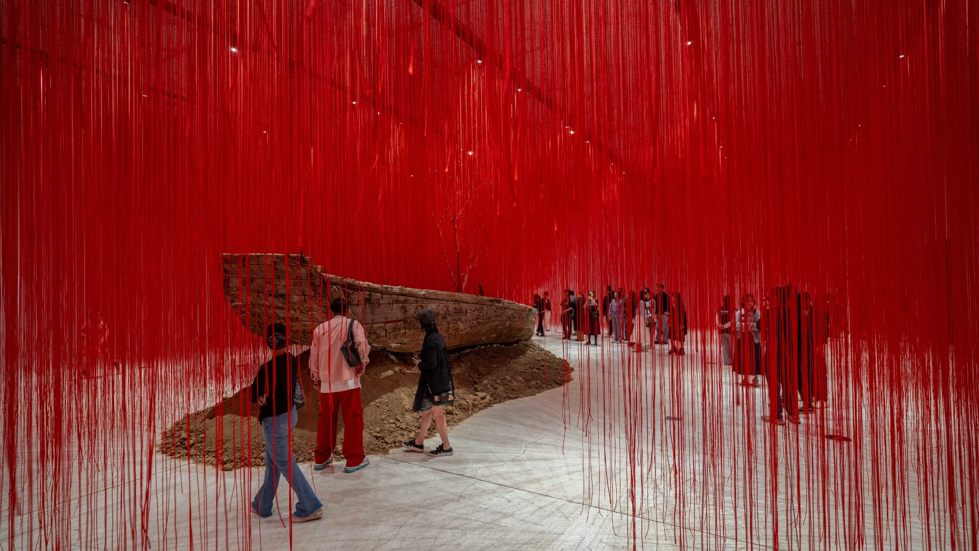 red string drapes around a wooden boat atop a pile of soil while people walk nearby