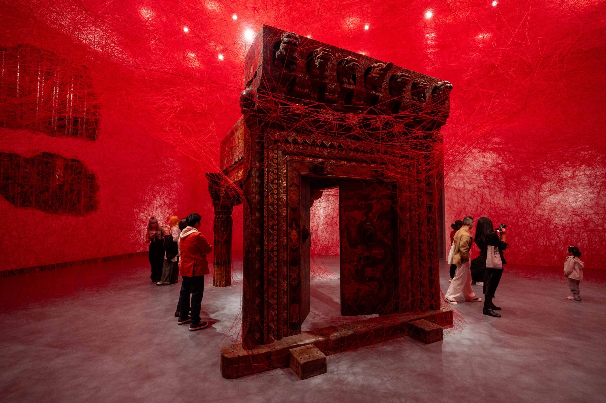 red string crisscrosses to wrap an elaborately carved structure while viewers walk around it