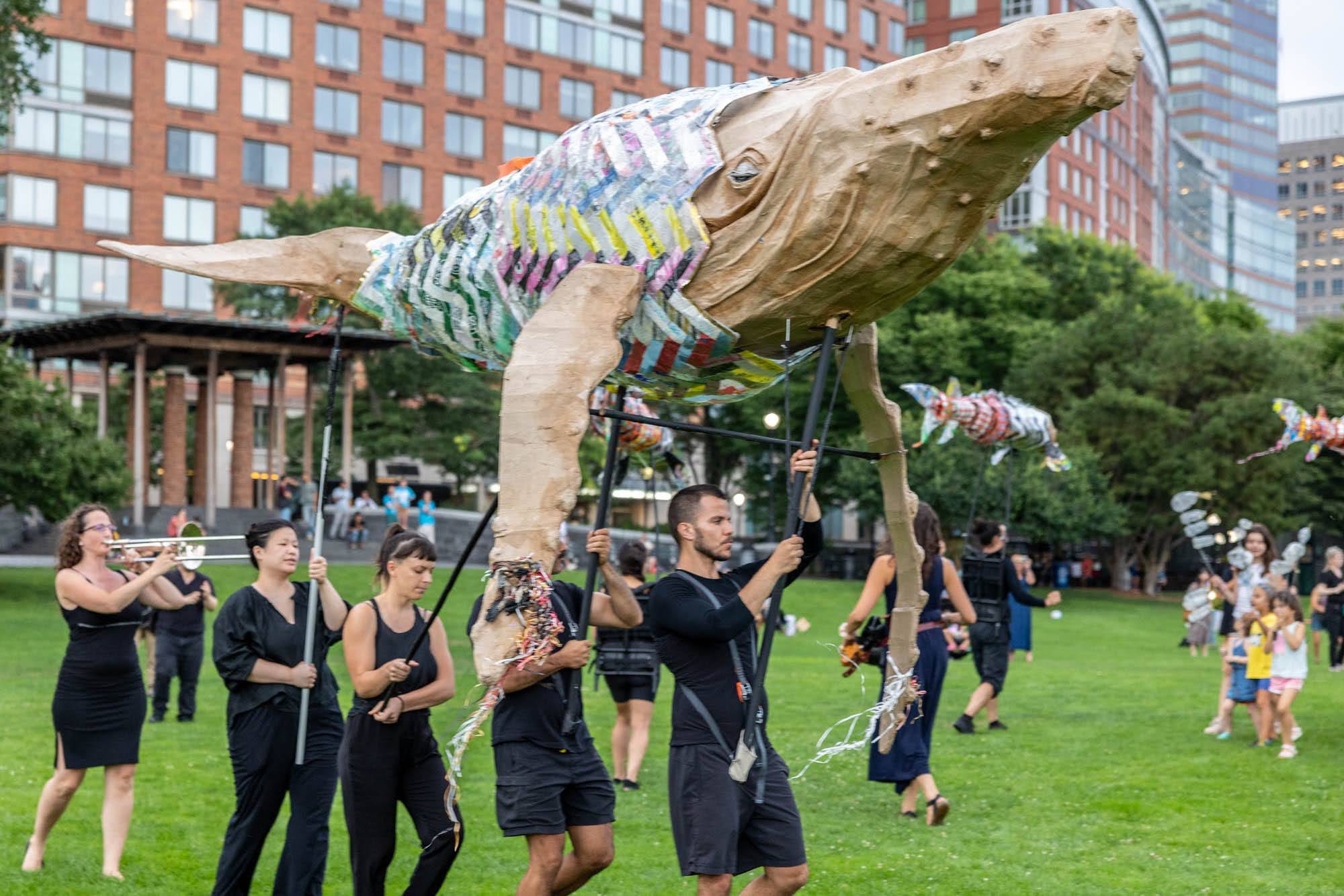people carry a large puppet of a whale on a pole