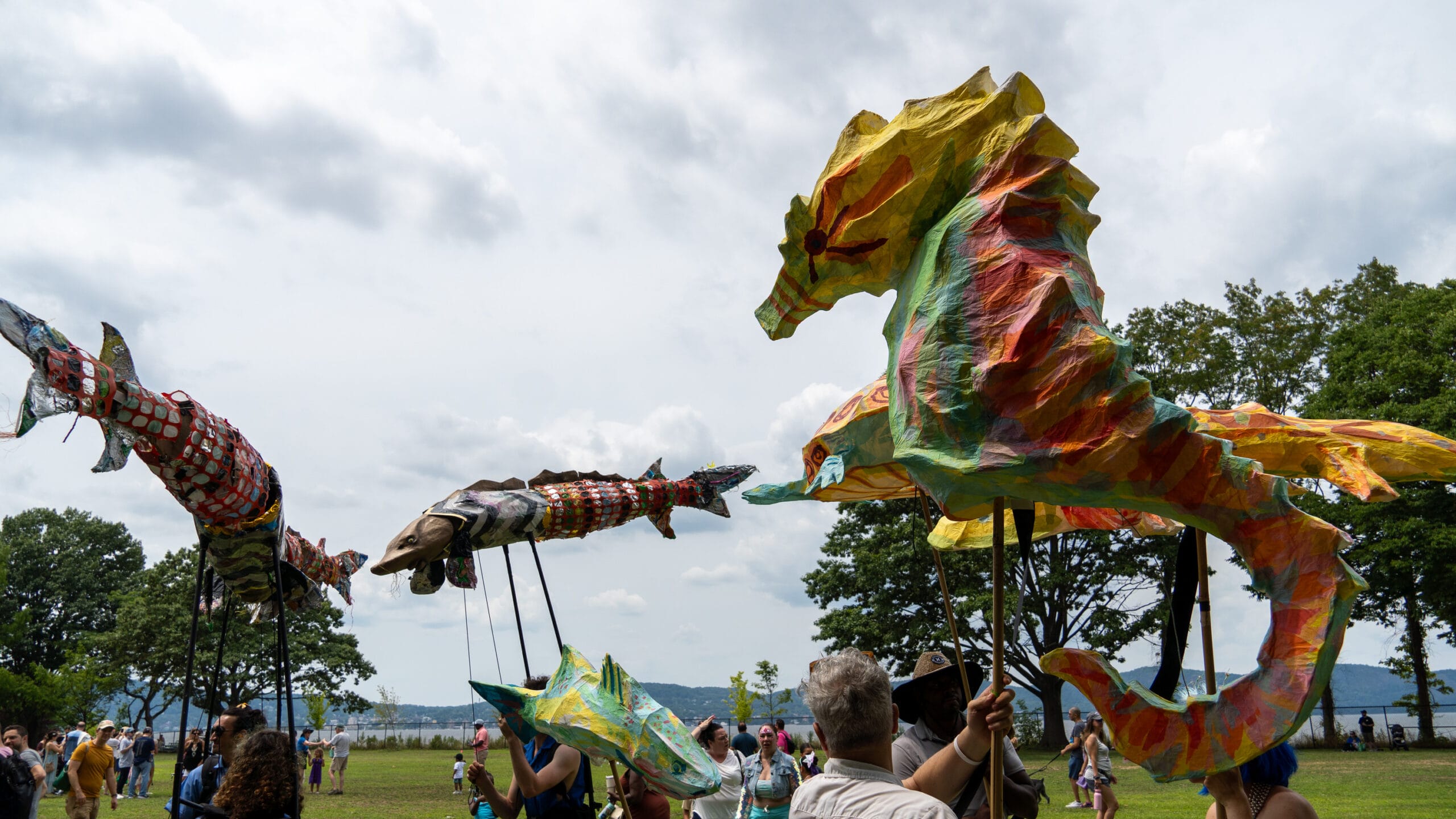a procession of people walk along a river carrying puppets of sturgeons and a seahorse on poles