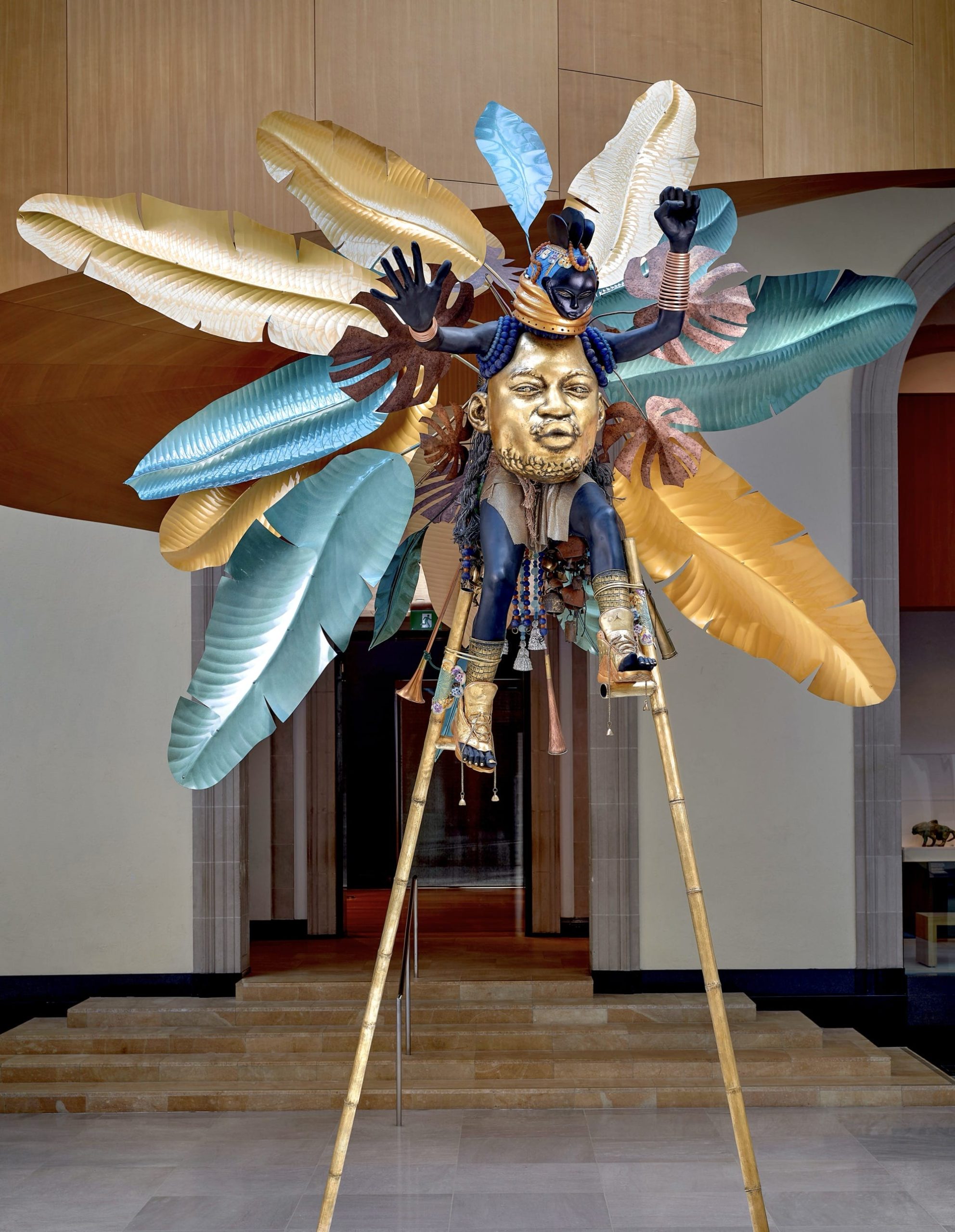 a vibrant sculpture with a gilded Black man's face in the center, serving as the body of another smaller, masked, dancing figure, surrounded by tropical leaves