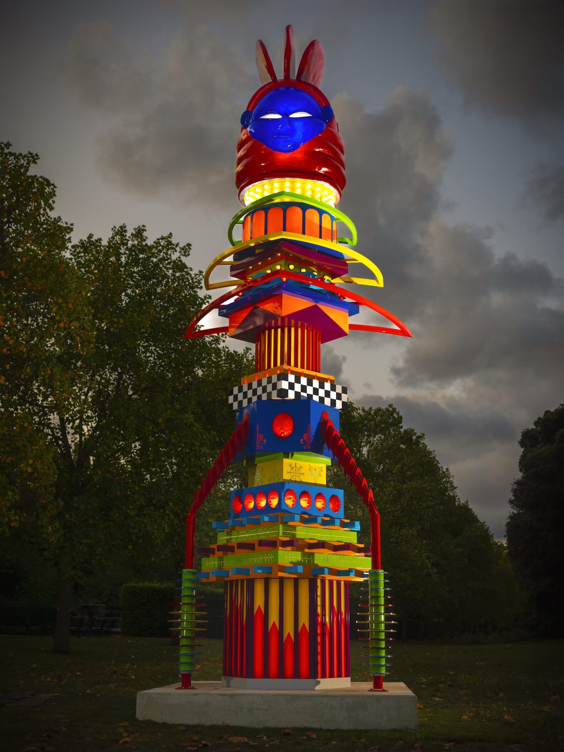 a tall, totemic, colorful sculpture illuminated at night, with a blue African mask on the top