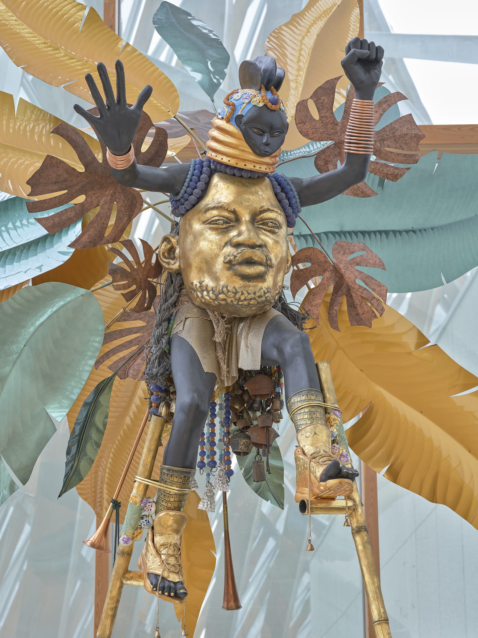 a detail of a vibrant sculpture with a gilded Black man's face in the center, serving as the body of another smaller, masked, dancing figure, surrounded by tropical leaves